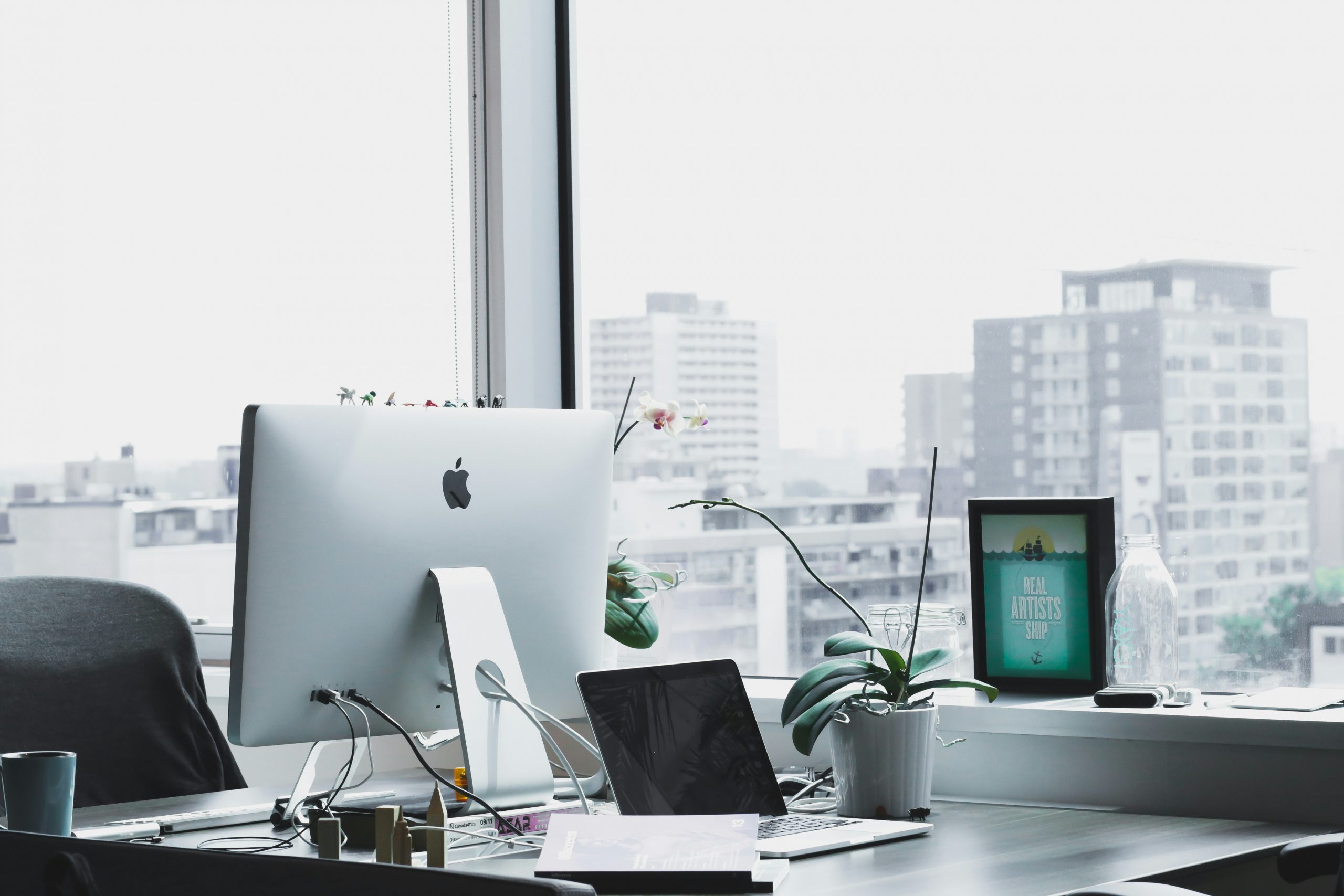 Modern office desk with Apple computer, laptop, plants, and city skyline in the background.